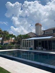 A modern house with large windows and a covered patio sits behind a rectangular swimming pool, surrounded by a stone deck. Palm trees and blue sky with fluffy clouds are in the background.