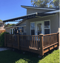 A person stands on a wooden deck attached to a beige house with white-framed glass doors. An extended black and white striped awning provides shade over the deck, and the yard is green and sunny.