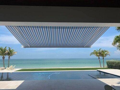 A striped retractable awning provides shade over a patio with a pool, overlooking a sandy beach and calm blue ocean, with palm trees framing the peaceful tropical scene.