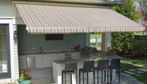 Outdoor kitchen with a striped retractable awning, bar seating for four, countertop, built-in grill, sink, and plants on a stone patio. Green lawn and trees are visible in the background.