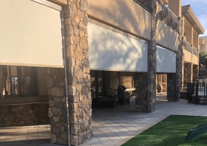 A building with stone pillars and large white outdoor blinds partially covering a tiled patio area, with artificial grass visible in the foreground.
