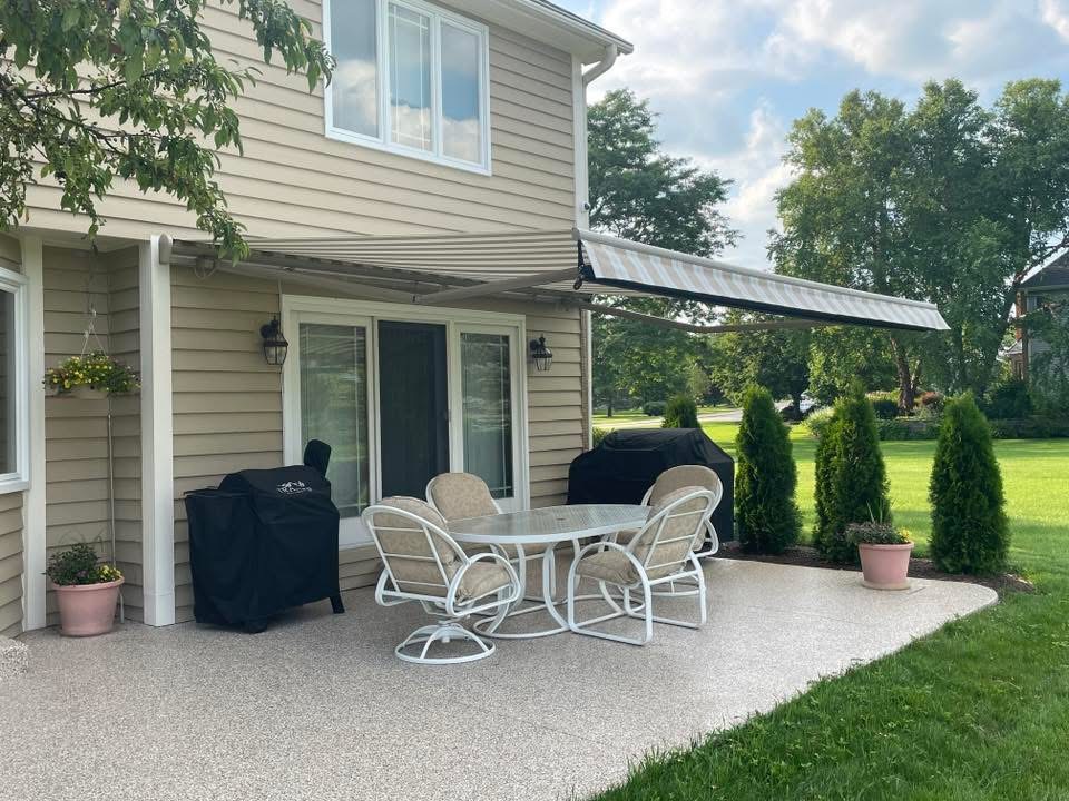 A backyard patio with a round table and four chairs, two covered grills, potted plants, and a striped retractable awning attached to a beige house. Green lawn and trees are visible in the background.