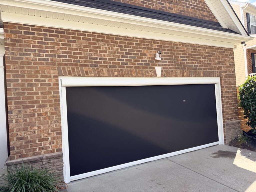 A closed black garage door with a white frame on a brick house, with concrete driveway in front and some green plants at the left and right edges.