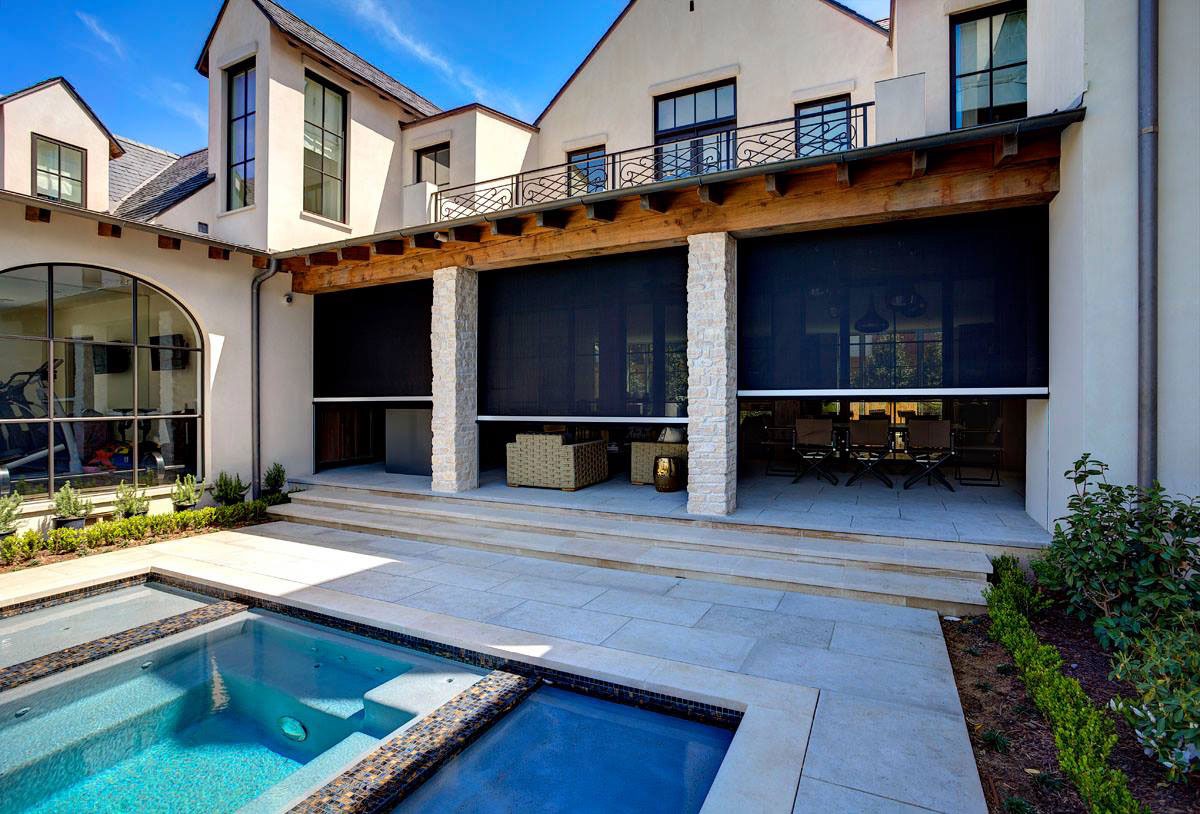 A modern house with a covered patio, outdoor seating, and dining areas behind retractable black screens. In front, there is a small pool and hot tub surrounded by tiled decking and landscaping.