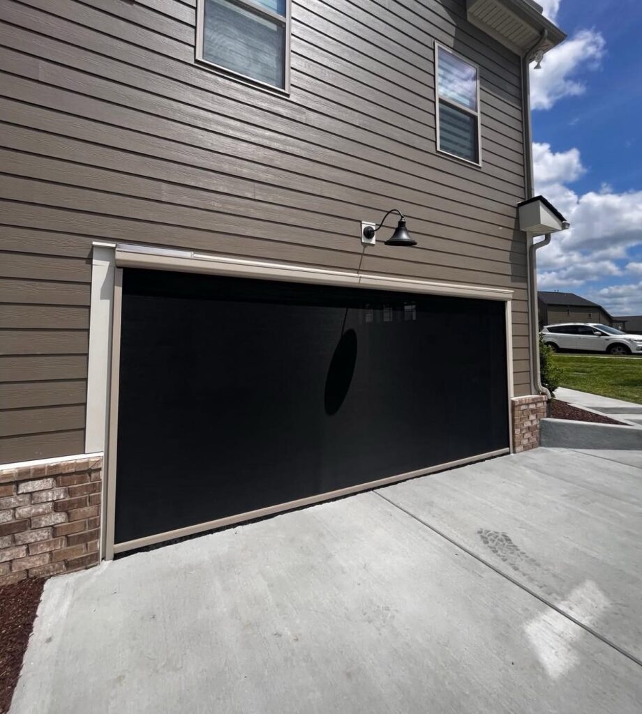A closed garage door with a black screen is attached to a beige house with brown siding and brick trim. There is an outdoor light above the garage and a clear blue sky with clouds in the background.