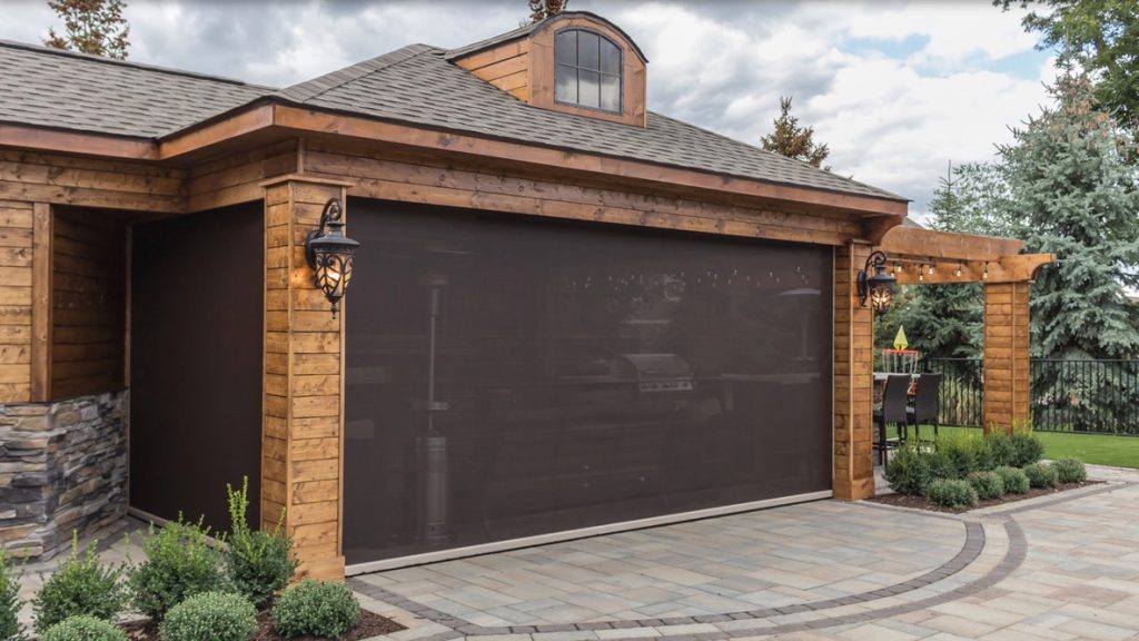 A wooden garage with stone accents features a closed, see-through dark screen door. Lantern-style lights hang on each side, with landscaped shrubs and a paved driveway in front. Trees and a grill are visible in the background.