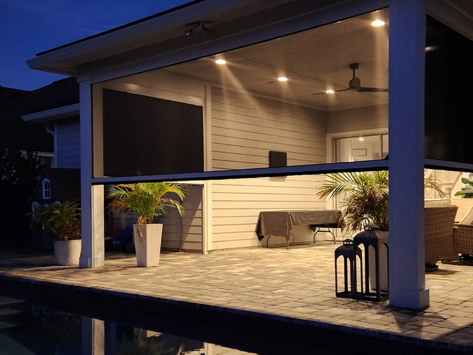 A covered patio at dusk with ceiling lights, potted plants, outdoor furniture, and a tiled floor next to a pool. The patio is partially enclosed with Sunpro motorized screen.
