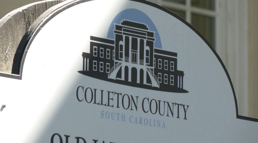 A close-up of a sign featuring an illustration of a courthouse building and the text Colleton County, South Carolina in black and blue letters.