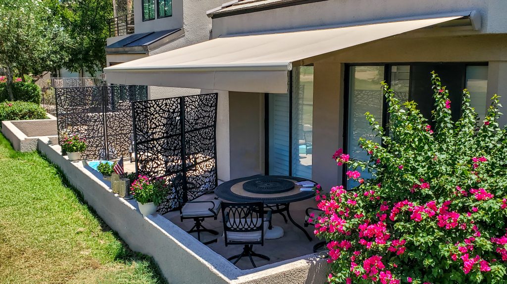 A cozy patio features a retractable awning shading a round table with four chairs, decorative privacy screens, lush potted plants, and bright pink flowers—all attached to a modern house.