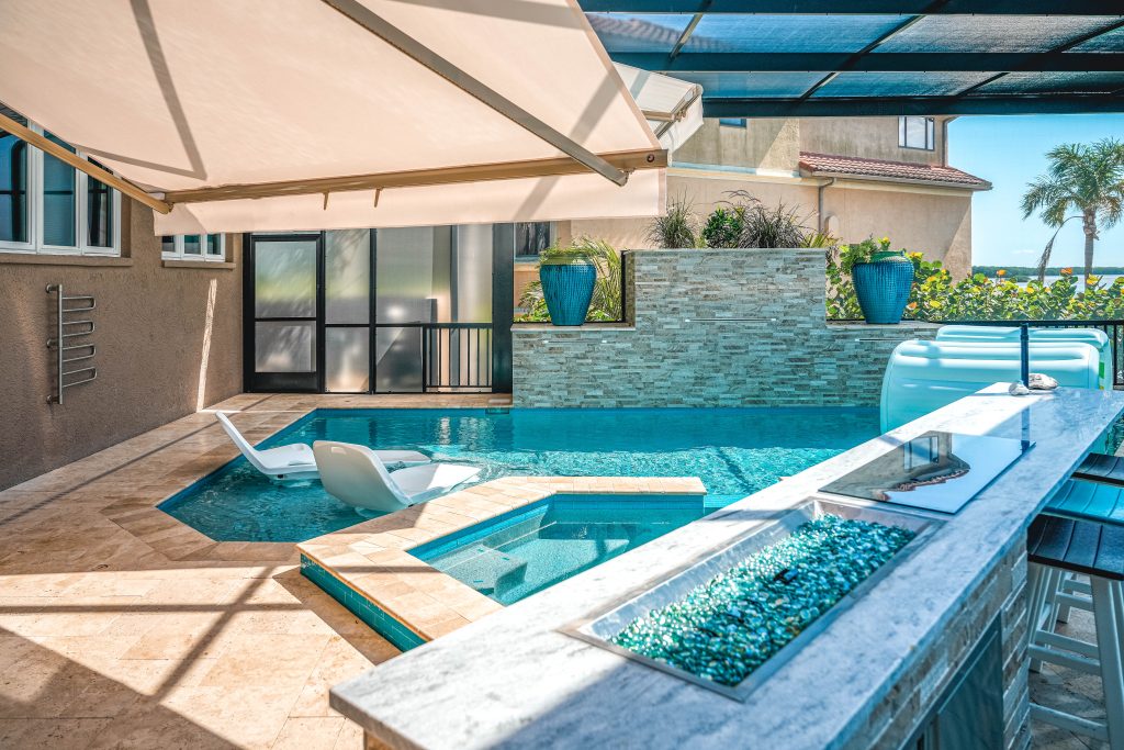 Modern backyard pool area with a built-in hot tub, lounge chairs in the water, a shaded bar with stools, glass fire pit, potted plants, and a view of palm trees and neighboring house under a clear sky.