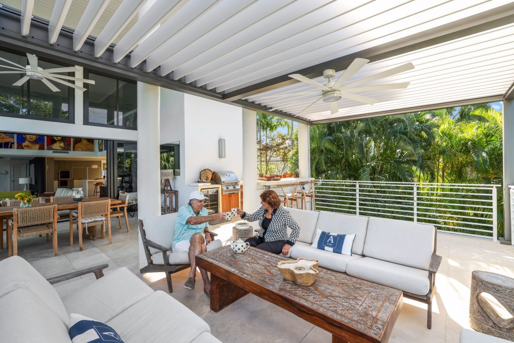 Two people sit and chat on a covered outdoor patio with white sofas, a wooden coffee table, ceiling fans, and tropical plants in the background. The space is bright and open with modern decor.