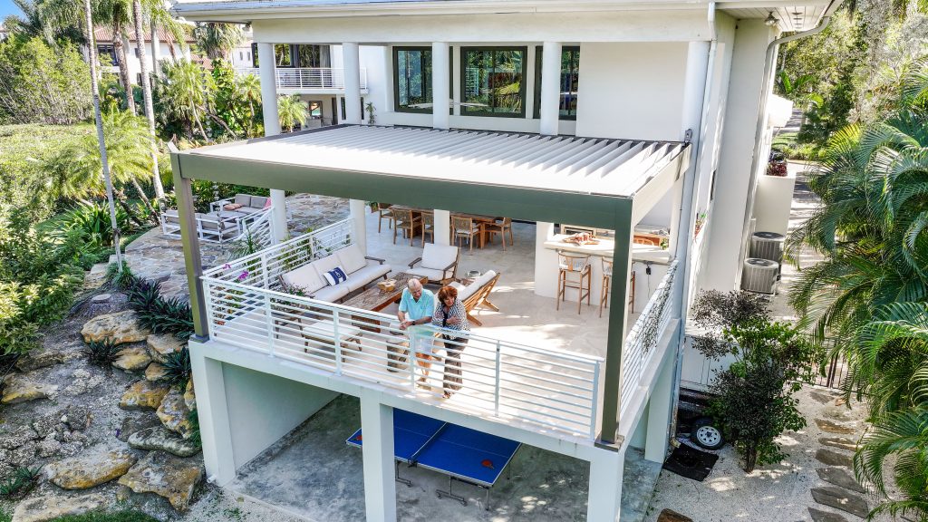 A couple stands on the upper balcony of a modern two-story house surrounded by lush greenery. Below, a spacious patio featuring outdoor dining furniture, a blue ping pong table, and a sleek pergola Azenco creates an inviting outdoor retreat.