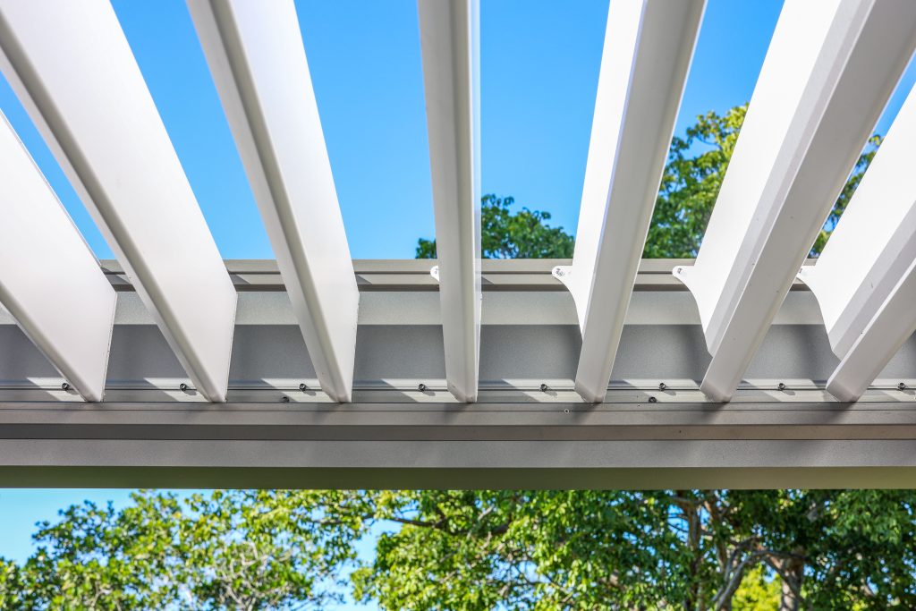 Close-up view looking up through the slats of a modern white pergola roof, with a clear blue sky and green trees visible in the background.