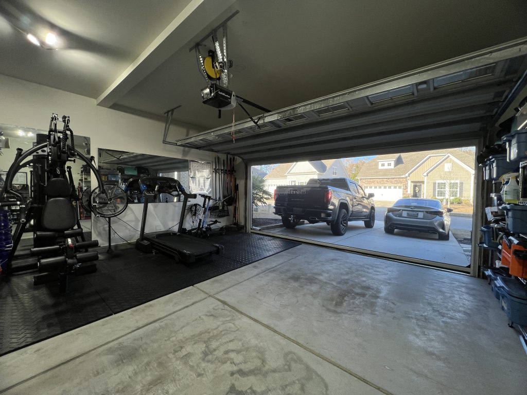 A spacious garage with gym equipment, including a treadmill and weight machine, sits next to an open garage door. Two cars are parked in the driveway outside a suburban house. Shelves line the right wall.