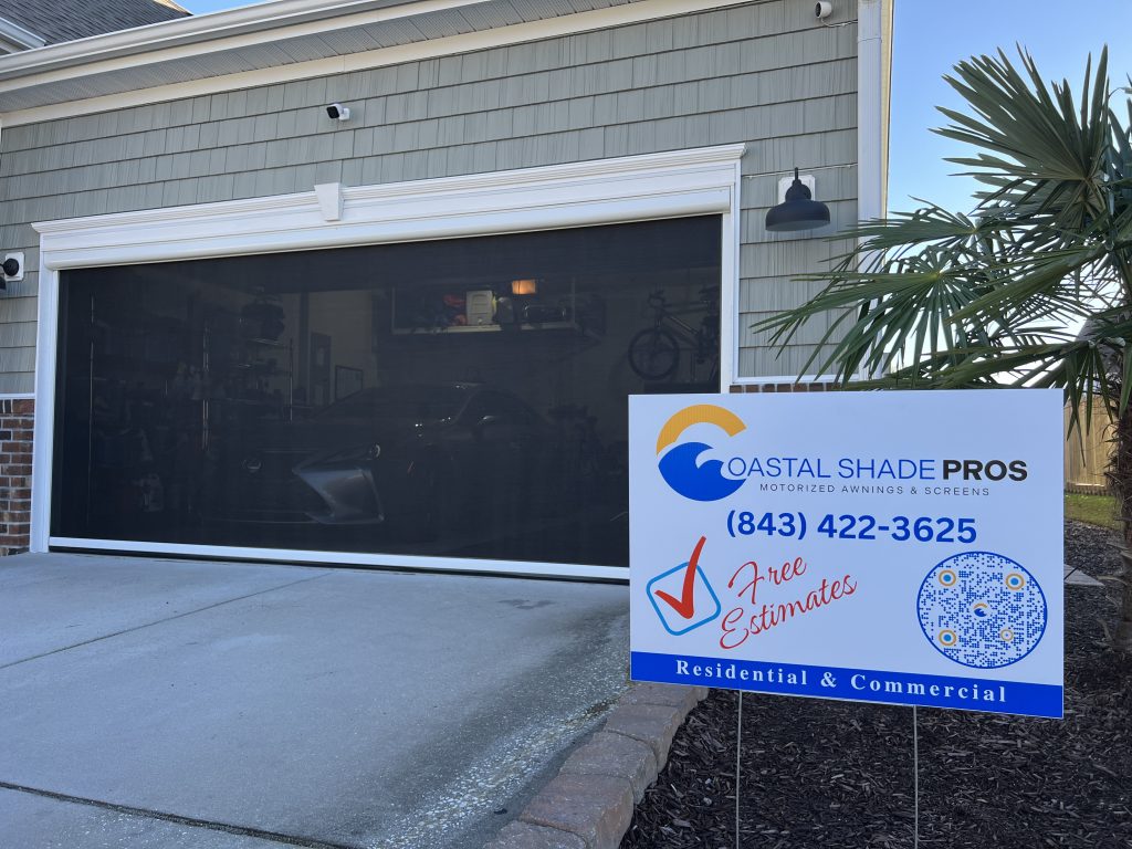 A garage with a black motorized screen is attached to a house. In front, a sign for Coastal Shade Pros displays their phone number, a checkmark for free estimates, and indicates residential and commercial services.