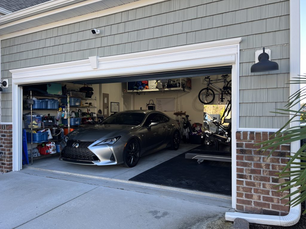 A gray sports car parked inside a two-car garage with various items stored on shelves, a bicycle hanging on the wall, and assorted tools and boxes around the space.