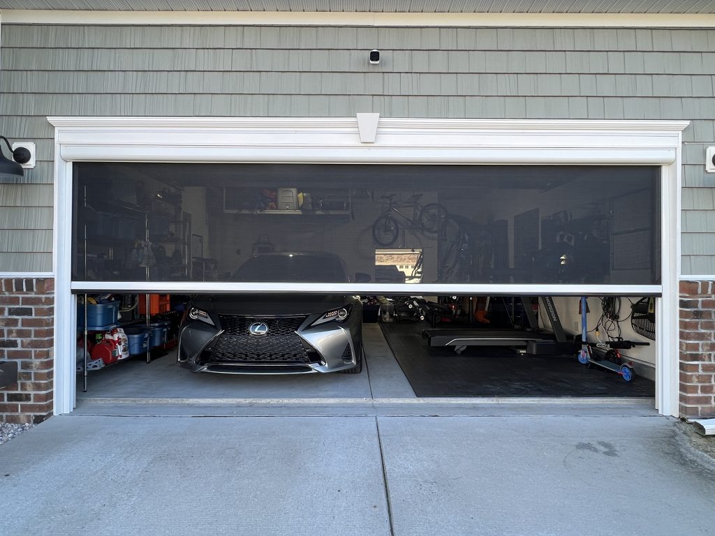 A partially open garage door reveals a silver Lexus car parked inside, with shelves of storage bins and various items visible on both sides and bicycles hanging on the back wall.