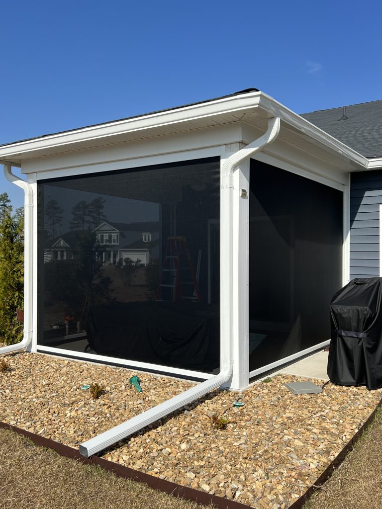 A screened-in porch with white framing attached to a house, surrounded by rocks and landscaping. A downspout extends from the gutter to the ground, and a grill is on the patio beside the porch. A house is visible in the background.