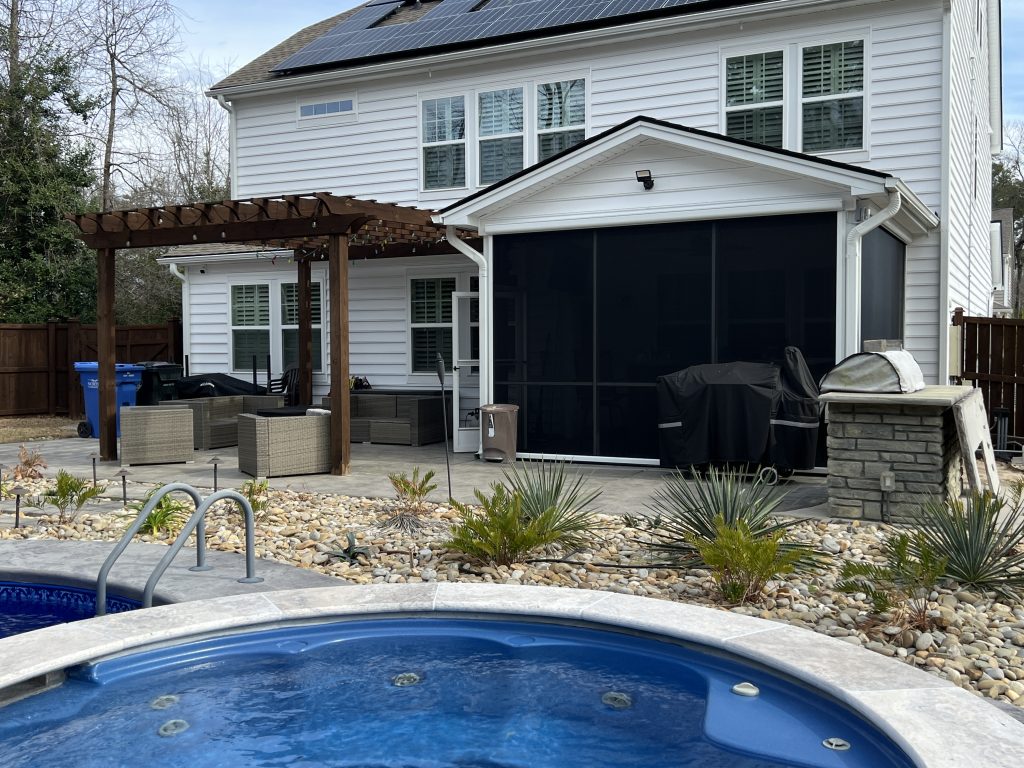 A backyard with a hot tub in the foreground, outdoor seating under a pergola, a grill, and a screened porch attached to a white two-story house with solar panels on the roof.