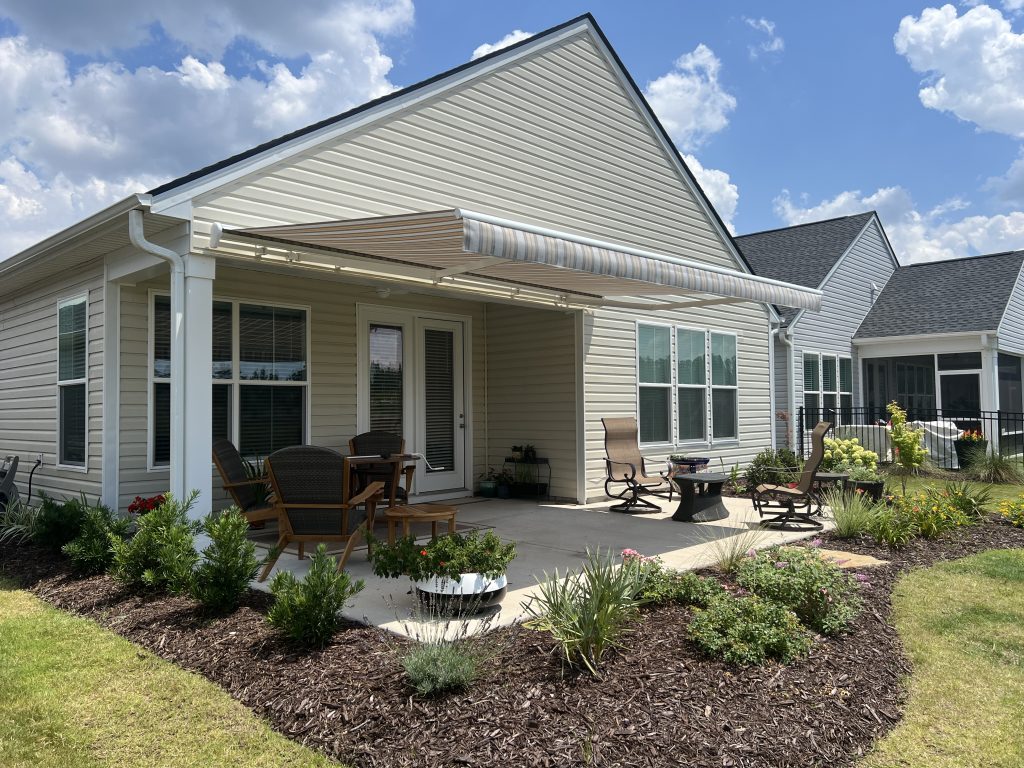 A beige house with a striped retractable awning shading a patio. The patio has outdoor chairs, a loveseat, and a fire pit, surrounded by landscaped flowerbeds and green grass under a partly cloudy sky.