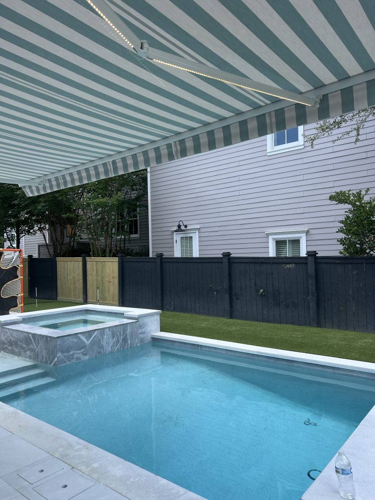 A backyard pool with clear blue water and an attached hot tub under a striped retractable awning. There is a black fence, green lawn, and a gray house in the background. A water bottle is on the pool deck.
