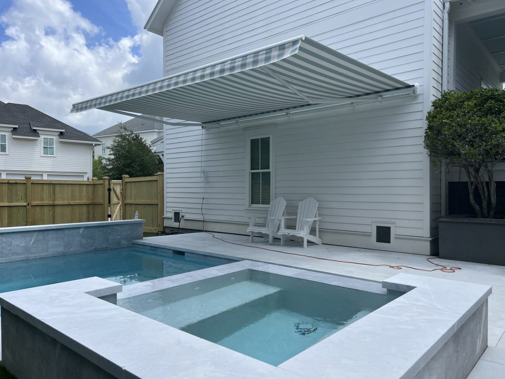 A backyard with a small pool and attached hot tub beside a white house. Two white Adirondack chairs sit under a green-and-white striped retractable awning. A wooden fence and some trees are in the background.