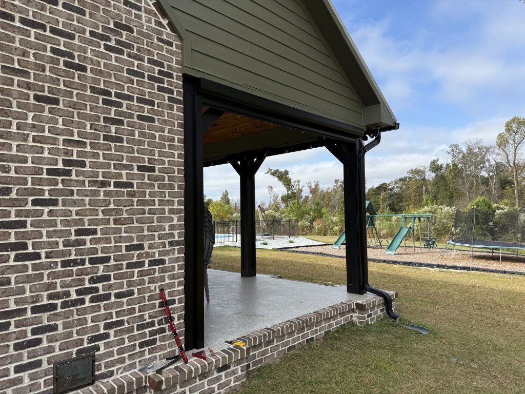 A brick pavilion with black metal supports opens onto a grassy area. In the background, there’s a playground with green swings and slides, and some trees under a partly cloudy sky.