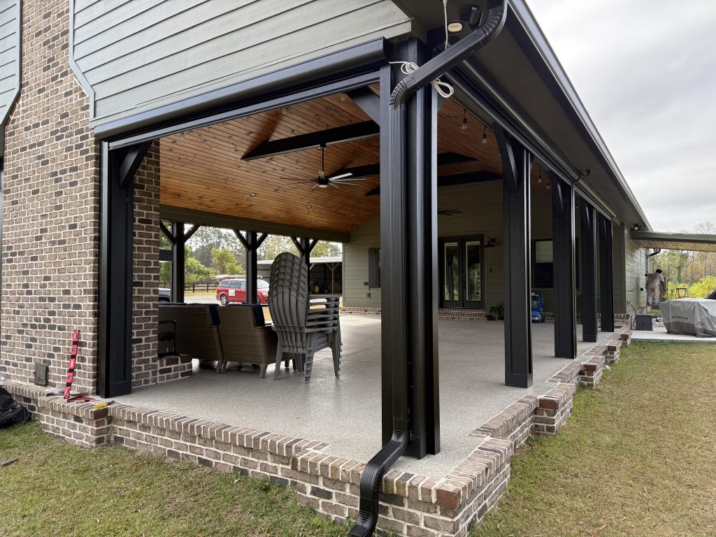 A covered patio with a brick foundation, black columns, and a wooden ceiling features outdoor furniture, a ceiling fan, and open space leading to a grassy yard.