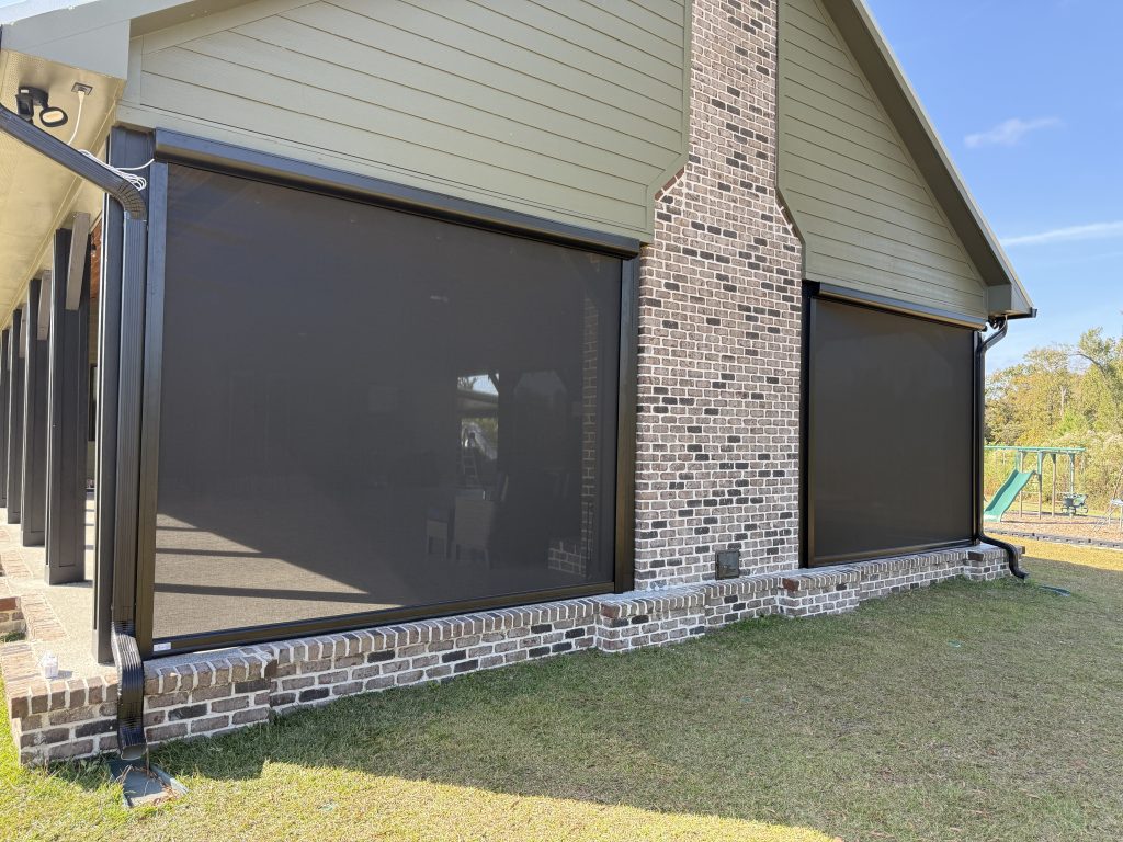 A house exterior with large, dark mesh screens covering the patio openings, framed by brick walls and siding, with grass and a glimpse of a playground structure in the background.
