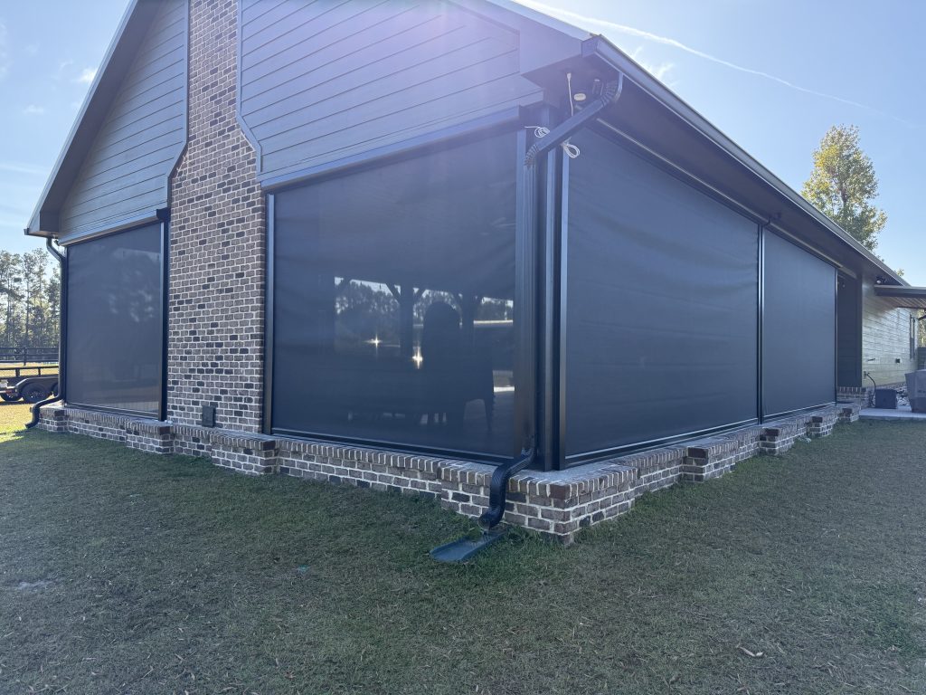 A brick house with large, dark retractable outdoor screens covering the windows and patio area, providing shade and privacy. The yard is grassy, and the weather is sunny with a clear sky.
