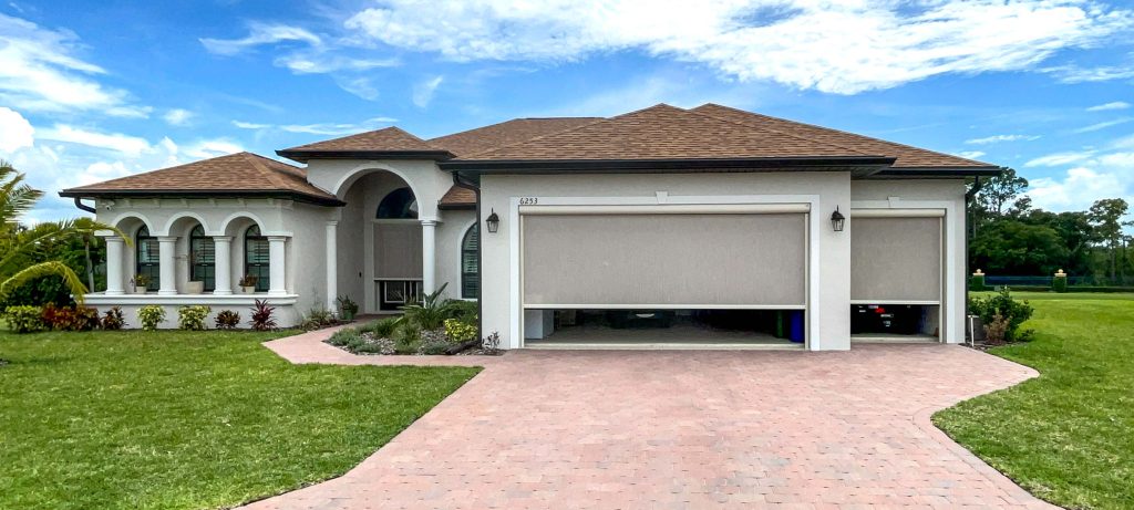 A modern single-story house with a brown roof, light-colored exterior, three-car garage, arched windows, a manicured lawn, and a brick driveway under a partly cloudy blue sky.