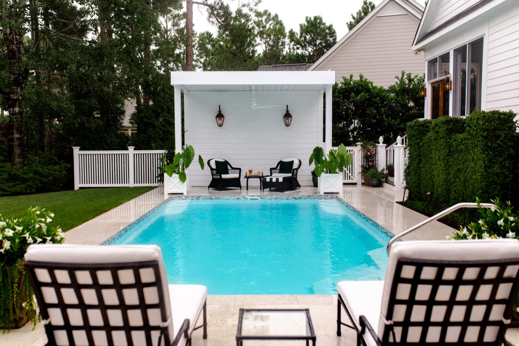 A backyard pool with two lounge chairs in the foreground, facing a covered patio with black chairs, a table, hanging lanterns, and lush greenery surrounding the area.