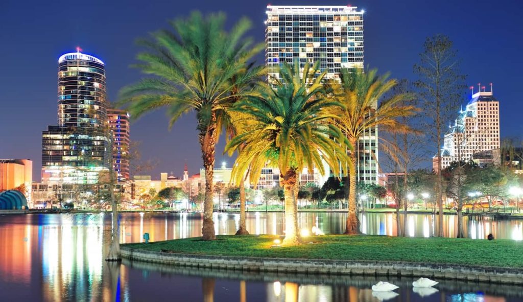 Palm trees on a small island in a lake, with city skyscrapers and buildings illuminated at night in the background; reflections shimmer on the water’s surface, creating a scene as relaxing as evenings under retractable awnings in Spring Hill FL.