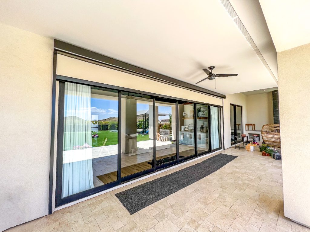 Covered patio with ceiling fan, beige tiled floor, and large glass sliding doors reflecting outdoor scenery of green hills and blue sky. Indoor furniture and decor are visible through the windows.