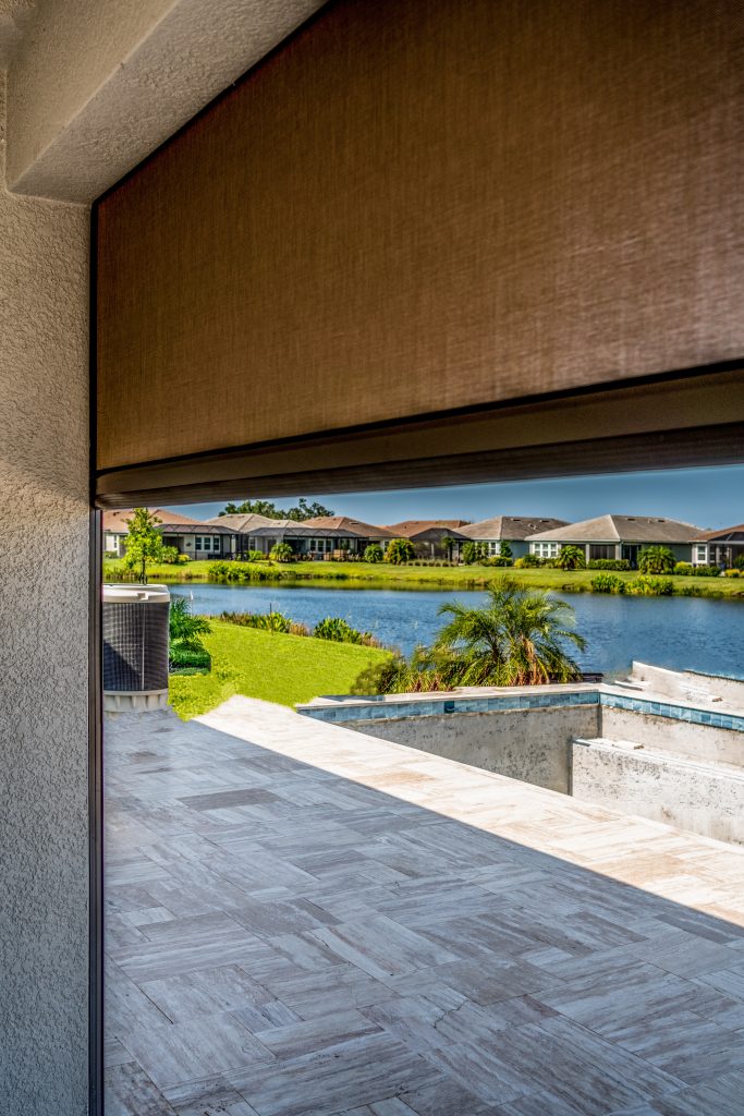 View from a covered patio with a brown sunshade partially down, overlooking a backyard with a pool under construction, green lawn, a small lake, and houses in the distance under a clear sky.
