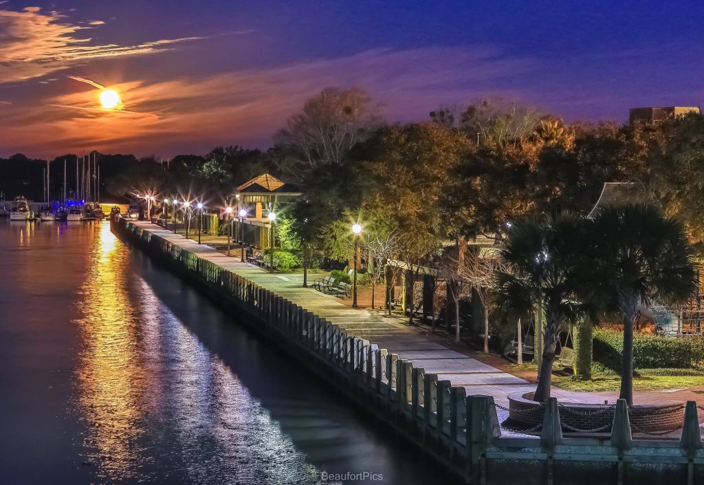 A vibrant sunset over a Beaufort County SC waterfront promenade lined with trees and lampposts, reflecting colorful lights on the calm water, with boats docked in the background.