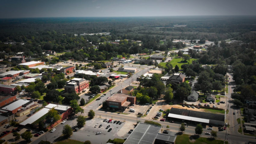 Aerial view of a small town with tree-lined streets, brick buildings, parking lots, and green spaces, surrounded by dense forest under a hazy sky.