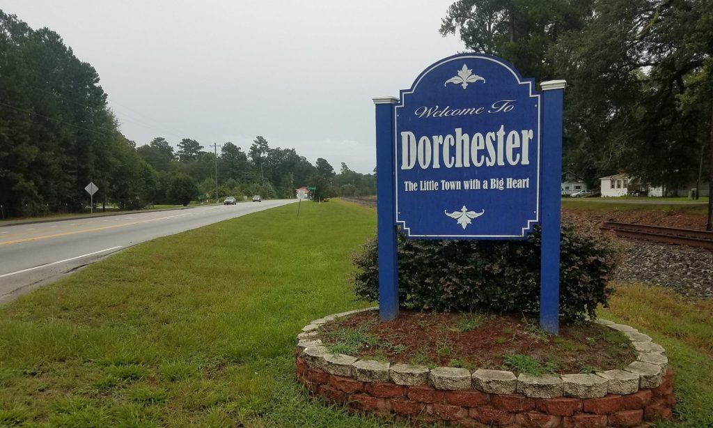 A blue roadside sign reads Welcome to Dorchester, The Little Town with a Big Heart. It stands beside a grassy area near a road and railroad tracks, with trees in the background.