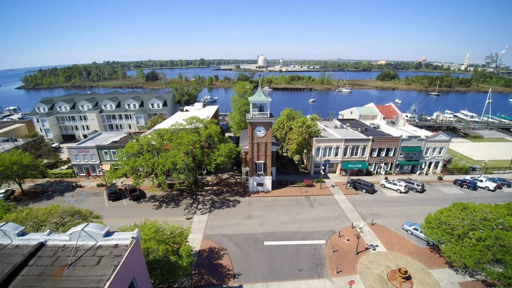 Aerial view of a small waterfront town with a clock tower, tree-lined streets, colorful buildings, parked cars, and a river with boats and green islands in the background on a sunny day.