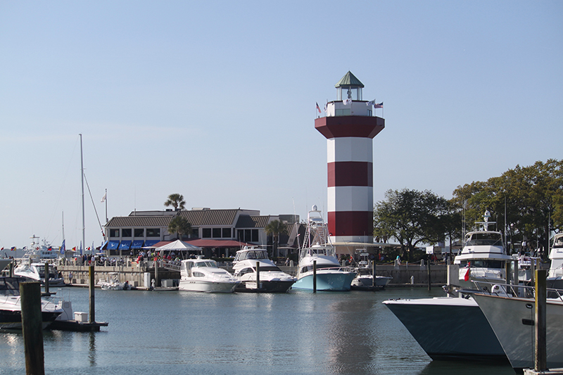 A marina with several docked boats sits beside a red-and-white striped lighthouse, surrounded by trees and buildings under a clear blue sky.