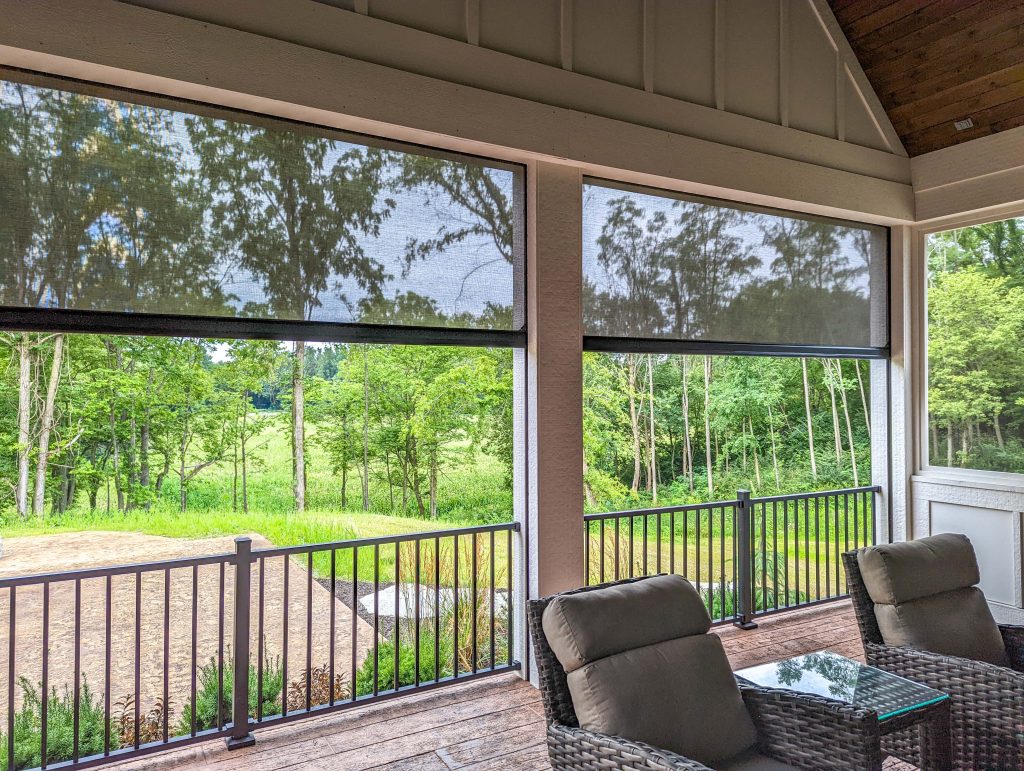 Outdoor patio area with wicker chairs and a glass coffee table, enclosed by black railing and mesh screen shades, overlooking a green landscape with trees and grass.