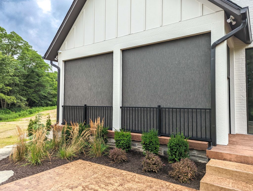 A modern house features two large outdoor window screens, black railings, white brick walls, tan steps, and a landscaped area with shrubs and ornamental grasses in the foreground.