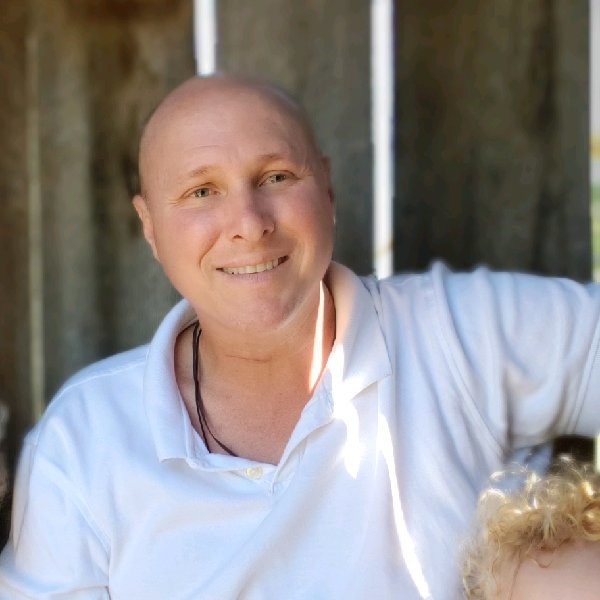 The Boval Family Chris Irminger in a white collared shirt relaxes beneath motorized awnings in a shaded outdoor area in Central Florida, with a blurred background. A person with curly blonde hair is partially visible in the lower right corner.