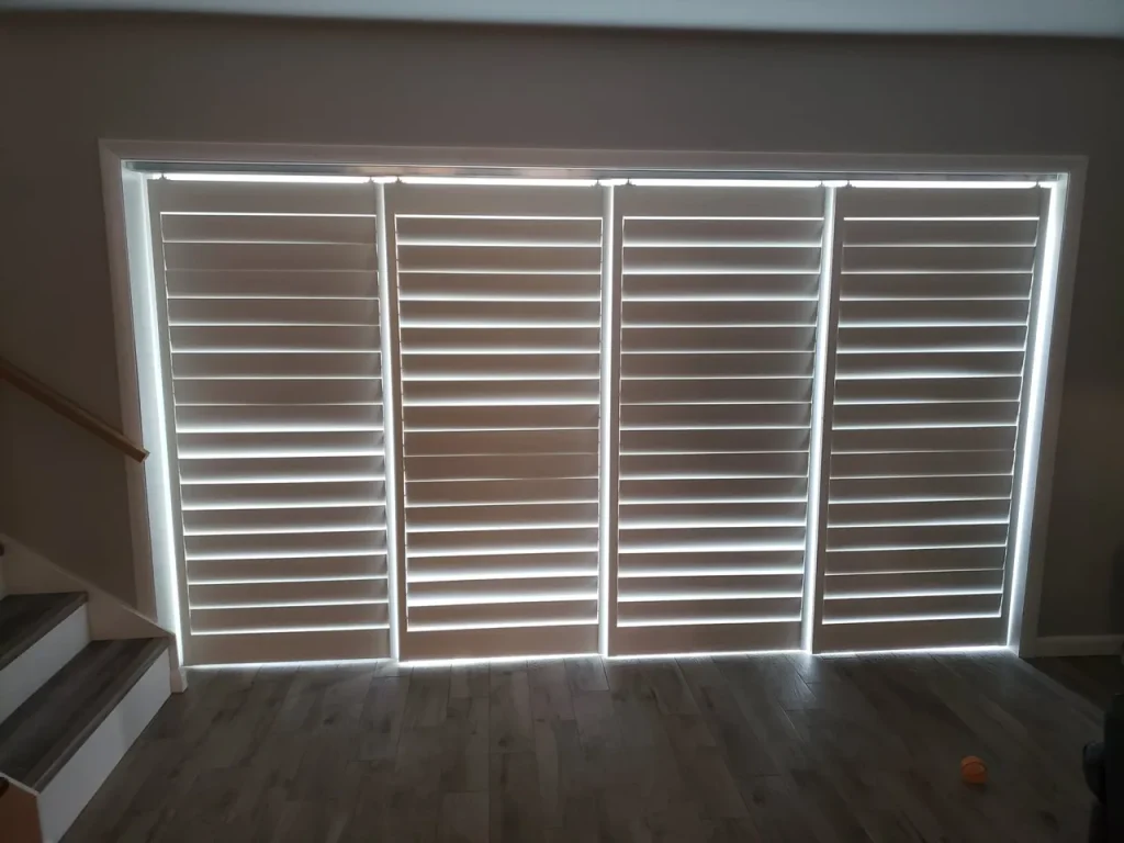 Four large window shutters are closed, with light streaming through the horizontal gaps between the slats, creating bright lines. The floor is wooden, and stairs are visible on the left side of the image.