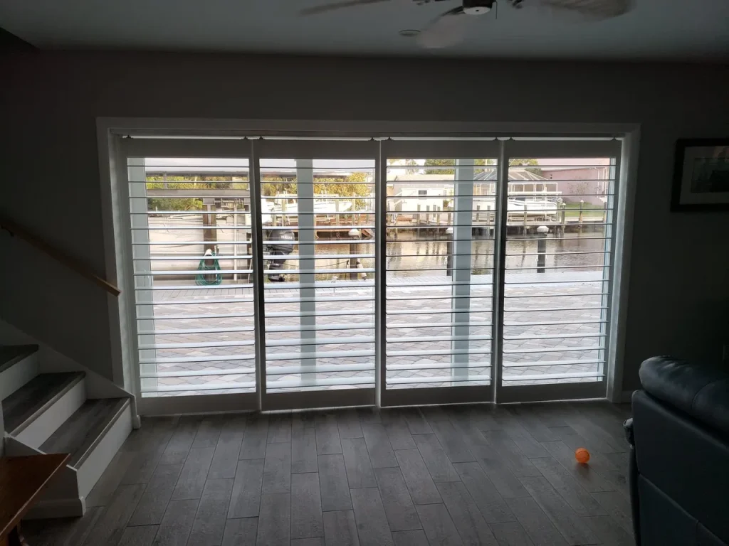 Large floor-to-ceiling glass doors with white blinds overlook a waterfront dock area with boats. Sunlight filters through the blinds, illuminating the hardwood floor inside the room. Steps and a couch are partially visible.