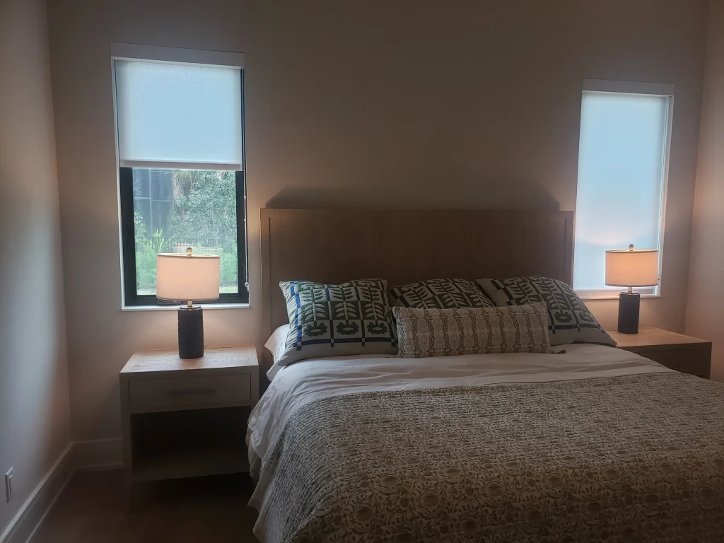 A bedroom with a neatly made bed, patterned pillows, and a textured blanket, flanked by two nightstands with matching lamps. Two narrow windows with shades let in natural light on either side of the bed.