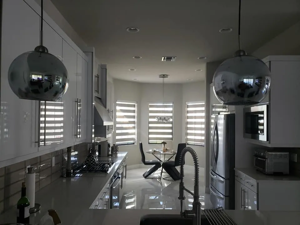 Modern kitchen with glossy white cabinets, stainless steel appliances, pendant lights, and a dining area with a round glass table and black chairs near large windows with striped blinds letting in sunlight.