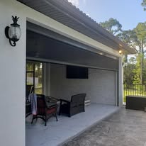 Covered outdoor patio with a seating area that includes chairs and a table. There is a wall-mounted TV and a sconce light on the white exterior wall. The area overlooks a fenced grassy yard with trees in the background.